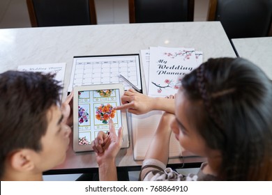 Over Shoulder View Of Young Asian Couple Choosing Bridal Bouquet With Help Of Digital Tablet While Planning Wedding Day At Home