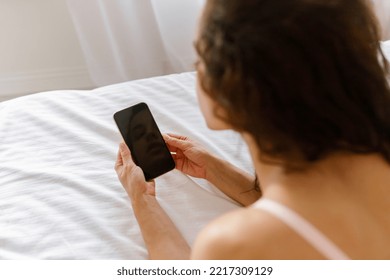 Over Shoulder View Of Young Brunette Woman In Bra Resting On Bed Indoors In The Morning Holding In Hands Smartphone Looking At Device Screen. Close Up Of Mobile Phone With Black Display In Female Hand