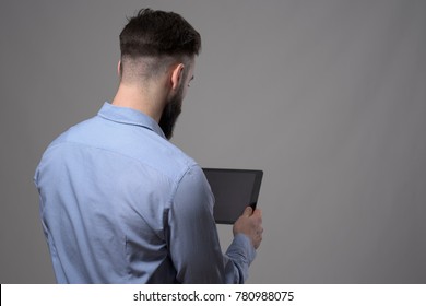 Over The Shoulder View Of Young Business Man Holding And Reading Blank Tablet Computer Over Gray Studio Background With Copyspace