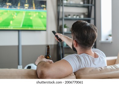 Over The Shoulder View Of A Young Man Spending His Day-off At Home Sitting On Sofa Watching Soccer Play Live Braodcast On Tv, Drinking Beer And Cheering For His Favourite Team. 