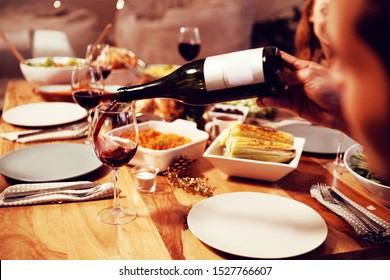 Over The Shoulder View Of A Young Mixed Race Man Sitting At A Table At Home Set For Thanksgiving Dinner With Friends Pouring A Glass Of Red Wine From A Bottle