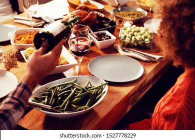 Over The Shoulder View Of A Young Mixed Race Man Sitting At A Table At Home Set For Thanksgiving Dinner Pouring A Glass Of Red Wine From A Bottle For A Young Mixed Race Woman Sitting At The Table