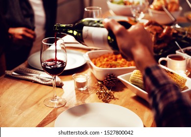 Over The Shoulder View Of A Young Mixed Race Man Sitting At A Table At Home Set For Thanksgiving Dinner Pouring A Glass Of Red Wine From A Bottle For A Man Sitting At The Table Opposite Him