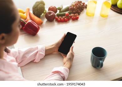 Over The Shoulder View Of Young Woman Holding And Using Mobile Phone With Black Empty Screen For Mockup Template, Cooking In Kitchen, Drinking Coffee In The Morning, Searching For Recipes Online