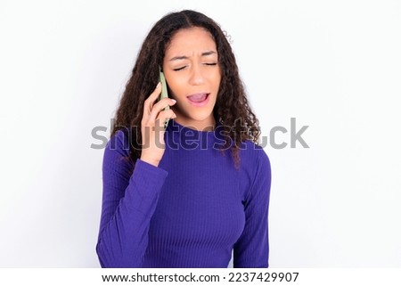 Overemotive happy beautiful teen girl wearing knitted purple sweater over white background laughs out positively hears funny story from friend during telephone conversation