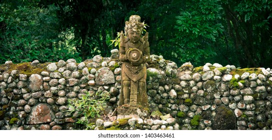 An overgrown ancient stone wall with Hindu statue amongst the trees in a tranquil forest of Bali, Indonesia.