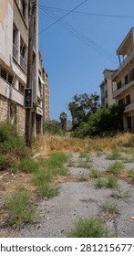 Overgrown path leads between weathered buildings. Grass sprouts from cracked asphalt, untraveled. Blue sky stretches overhead, wires cross the view. Sunlight bathes abandoned architecture, silent