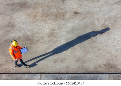 Overhead Aerial View Of Anonymous Construction Worker Or Builder Wearing A Hi Vis Jacket, A Hardhat And Carrying A Clipboard On A Building Site With Their Shadow Stretching Out In Front Of Them