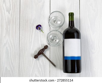 Overhead Angled View Of A Large Bottle Of Red Wine, Drinking Glasses, Flower And Antique Corkscrew On White Wooden Boards. 