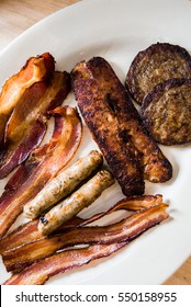 Overhead Of Browned Sausage Links And Patties With Crispy Bacon In Morning Light On White Oval Platter On Restaurant Table.