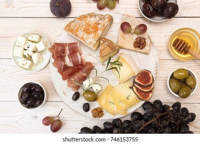 Overhead Of Cheese Plate With Pieces Moldy Cheese, Prosciutto, Pickled Plums, Olives, Grapes, Figs, Honey, Pear, Nuts On White Wooden Background. Top View. Copy Space.