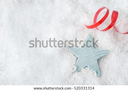 Overhead Christmas background shot.  A pale blue wooden star, resting in white artificial snow, with red swirl of ribbon above and copy space to the left.