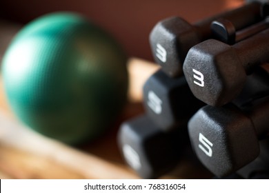 Overhead Close Up On A Rack Of Black Rubber Coated Dumbbell Weights, On A Wood Bench, With A Heavy Medicine Ball In The Blurry Background, At A Physical Therapy And Fitness Gym 