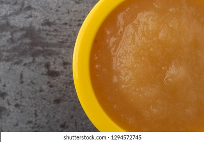 Overhead Close View Of A Small Yellow Bowl Filled With Applesauce On A Gray Background.