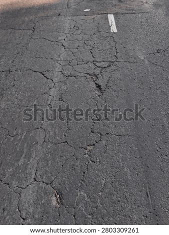 An overhead close-up of an empty asphalt road surface showing extensive cracking and rough textures. The image focuses on the natural patterns formed by wear and age, giving it an abstract, gritty visual feel.