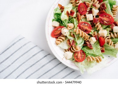 Overhead, Close-up Shot Of BLT Pasta Salad With Crispy Bacon, Grape Tomatoes, Green Leafy Lettuce And Fresh Mozzarella In White Bowl With Blue Striped Linen.