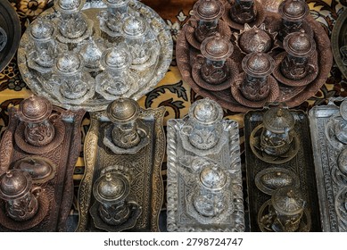 An overhead close-up shot captures multiple intricately designed silver and copper Turkish tea or coffee sets arranged on decorative metallic trays. These traditional metalware items, featuring ornate patterns and matching lids, saucers, and cups, are displayed on a colorful, patterned rug, likely in a bustling marketplace.