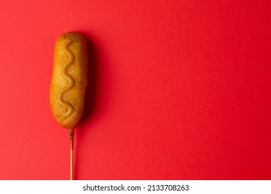 Overhead Close-up Shot Of Corn Dog With Mustered Sauce Over Skewer On Red Background With Copy Space. Unaltered, Copy Space, Unhealthy Food, Meat, Sausage And Fried Food.