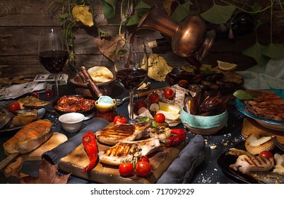 Overhead Of Dinner Table. Assorted Grilled Barbecue Meat And Seafood. Pouring Wine From Jar. Pork Steaks, Salmon Trout, Mussels, Shrimps, Dried Tomato, Cherry Tomato, Chili Pepper, Glasses Of Wine.