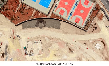 Overhead drone view capturing new vibrant basketball and tennis courts adjacent to a large urban construction site undergoing active development, representing growth and future infrastructure