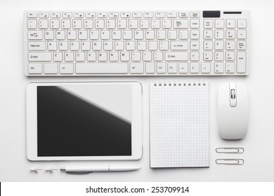 Overhead Of Essential Office Objects In Order On White Desk. Notebook, Computer Keyboard And Mouse, Tablet Pc, Pen, Push Pins, Paper Clips 