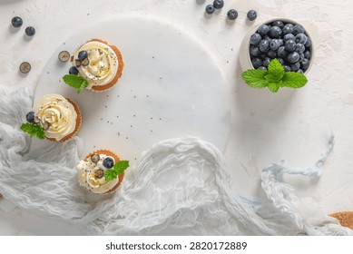 An overhead flat lay composition features three cupcakes with white frosting and a bowl of blueberries on a textured white surface.
