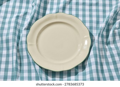 Overhead flat lay shot of an empty minimalist white ceramic plate centered on a rustic, wrinkled blue and white checkered linen tablecloth. Captured in Italy under bright, sunny summer daylight with soft shadows. Ideal as a food mockup backdrop, culinary template, or restaurant menu background