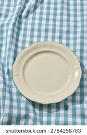 Overhead flat lay shot of an empty minimalist white ceramic plate placed on a rustic, wrinkled blue and white checkered linen tablecloth. Captured under bright, sunny summer daylight with soft, natural shadows highlighting the gingham fabric texture. Ideal as a food mockup backdrop, culinary template, restaurant menu background, or picnic concept layout