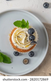 Overhead flat lay shot of a single cupcake with swirled white buttercream, fresh blueberries, and mint on a light blue ceramic plate, lit by soft natural light.