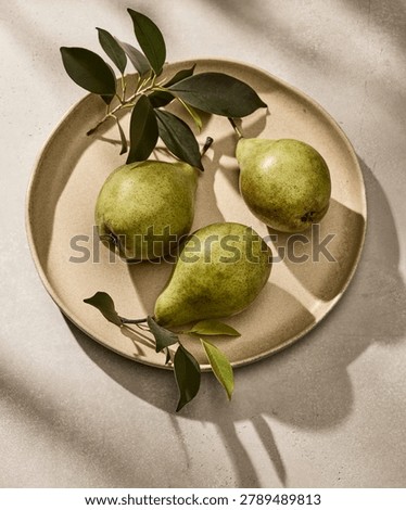 Overhead flat lay shot of three fresh, organic Italian pears with natural green leaves arranged beautifully on a rustic ceramic plate. The plate sits on a kitchen tabletop surface, captured in a warm, golden color palette under bright, sunny daylight with soft, elegant shadows. This harmonious, minimalist still life composition is perfect for kitchen wall art, restaurant posters, organic fruit branding, and autumn lifestyle print designs