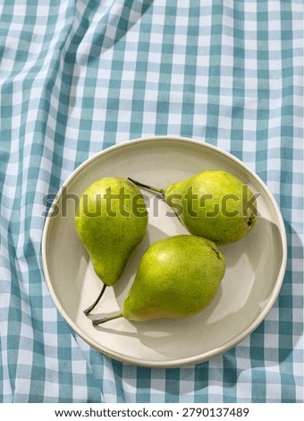 Overhead flat lay shot of three fresh organic green pears arranged on a light ceramic plate. The dish rests on a classic blue and white checkered gingham tablecloth background. Captured in bright, warm, sunny daylight casting beautiful natural shadows across the scene. Ideal for rustic kitchen art, traditional recipe blogs, summer or autumn food styling, and organic harvest marketing