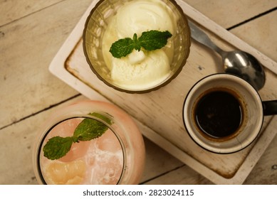 Overhead flat lay view of a cafe dessert set featuring scoops of creamy vanilla ice cream topped with fresh mint sprig and a hot shot of dark espresso in a ceramic cup on a white wooden tray, paired with a cold pink iced fruit drink in a glass on a rustic tabletop.