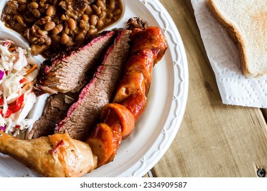 Overhead of a full plate of smoked chicken, sausage and barbecue beef brisket with baked beans and fresh coleslaw on wooden table with white bread on a paper napkin in the background. - Shutterstock ID 2334909587