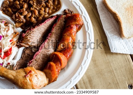 Overhead of a full plate of smoked chicken, sausage and barbecue beef brisket with baked beans and fresh coleslaw on wooden table with white bread on a paper napkin in the background.