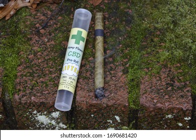 Overhead High Angle CBD Palm Pre-rolled Leaf Blunt Wrap, Hemp Flower, Lit Smoking, Green Mossy Corner Brick Step Outdoors Clear Plastic Labeled Cigar Tube 