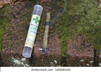 Overhead High Angle CBD Palm Pre-rolled Leaf Blunt Wrap, Hemp Flower, Lit Smoking, Green Mossy Corner Brick Step Outdoors Clear Plastic Labeled Cigar Tube 