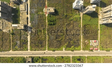 An overhead, high-angle, or drone shot captures a mix of undeveloped agricultural land or fields divided into rectangular plots, bordered by a main paved road and new high-rise residential or commercial buildings under construction and already built on the left and right sides. The landscape shows a clear transition from rural or undeveloped land to urban expansion, featuring some patches of green grass, brown soil, and muddy areas within the plots, indicating potential recent development or land preparation. The contrast between the grid-like fields and the modern structures highlights real estate development and urban sprawl.