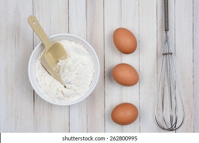 Overhead Image Of A Bowl Of Flour, Three Brown Eggs And A Metal Whisk On A Rustic Wooden Kitchen Table. 