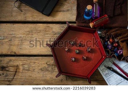 Overhead image a dice tray with role-playing gaming dice surrounded by TTRPG equipment on a wooden table.