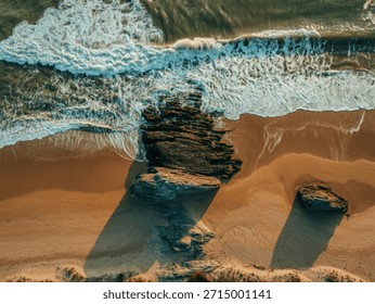 An overhead perspective captures dynamic ocean waves breaking against dark, rugged rocks and a sandy beach, showcasing natural coastal beauty.