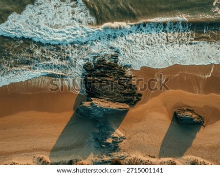 An overhead perspective captures dynamic ocean waves breaking against dark, rugged rocks and a sandy beach, showcasing natural coastal beauty.