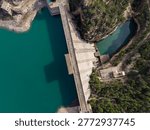 Overhead perspective of a large concrete dam separating a turquoise reservoir from a narrow canyon river gorge, surrounded by rocky cliffs and green Mediterranean vegetation