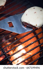 Overhead Perspective Of Polished Metal Spatula With Bottle Opener Removing Cheese Burger Patty With Slice Of Melted Pepper Jack Cheese On Barbecue Grate With Glowing Orange Charcoal Coals And Flames