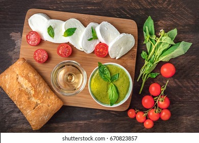 Overhead Photo Of Buffalo Mozzarella Cheese With Cherry Tomatoes, Green Basil Leaves, The Ingredients Of The Caprese Salad, A Glass Of White Wine, Pesto Sauce, And A Ciabatta, On A Dark Rustic Texture