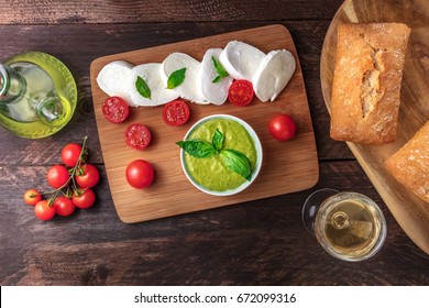 Overhead Photo Of Buffalo Mozzarella Cheese With Cherry Tomatoes, Basil Leaves, Ingredients Of Caprese Salad, Glass Of White Wine, Pesto Sauce, Olive Oil, Ciabattas, On Rustic Texture With Copy Space