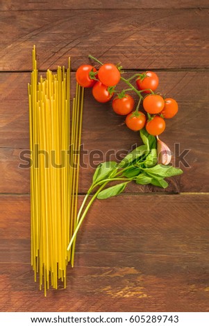 An overhead photo of a letter P for pasta, formed by spaghetti, cherry tomatoes, a garlic clove, and a sprig of basil leaves, on a dark wooden background texture with copy space