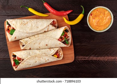 An overhead photo of Mexican burritos with beef, rice, black beans, and vegetables, with a cheese sauce, chili peppers, and a place for text, shot from above on dark wooden rustic textures