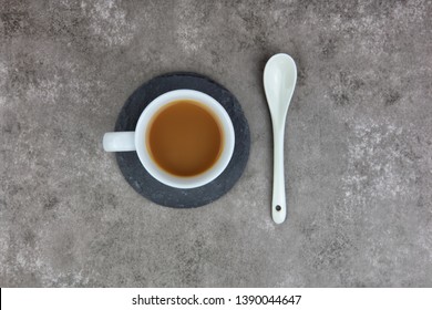 Overhead Photo Of Milky Tea In White Cup With Spoon On A Circular Slate Coaster On A Pale Tabletop