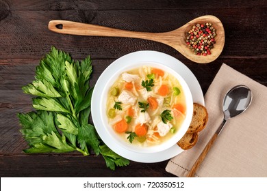 An Overhead Photo Of A Plate Of Chicken And Noodles Soup, Shot From Above On A Dark Rustic Texture With A Spoon, A Wooden Ladle With Peppercorns, Slices Of Bread, A Celery Branch, And A Place For Text