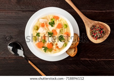 An overhead photo of a plate of chicken and noodles soup, shot from above on a dark rustic texture with a spoon, a wooden ladle with peppercorns, slices of bread, and a place for text